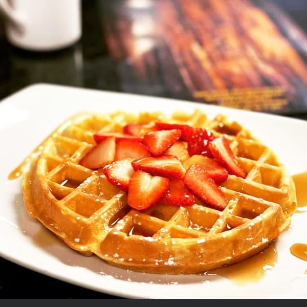 Golden waffle topped with sliced strawberries, powdered sugar, and maple syrup on a white plate.